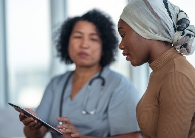 A young black woman with cancer is meeting with her female doctor of Asian descent. The patient is wearing a scarf on her head to hide her hair loss. The doctor is showing the woman test results on a tablet computer. The patient is listening to her doctor's advice regarding further treatment.
