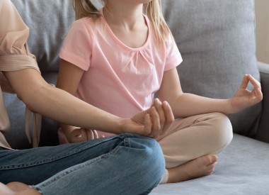 In daytime serene young mother and preschool cute daughter sitting in lotus position on couch at home closed eyes doing yoga relaxation exercise meditating, healthy habits lifestyle, keep calm concept