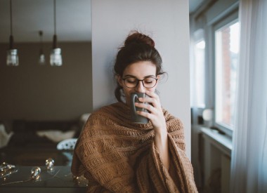 Photo of a young woman drinking her first cup of coffee early in the morning, enjoying her space and her freedom covered up in a blanket