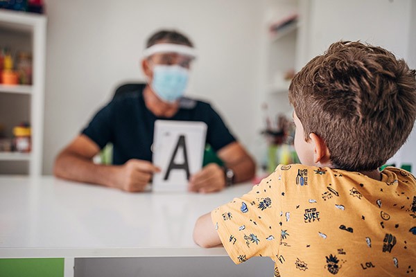 Two people, male speech therapist with surgical mask and protective visor teaching a boy to say letter A.