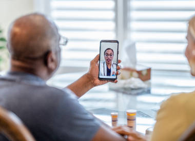 Remote medical visit between a patient and spouse and their female doctor from the patients point of view while they sit at their kitchen table.