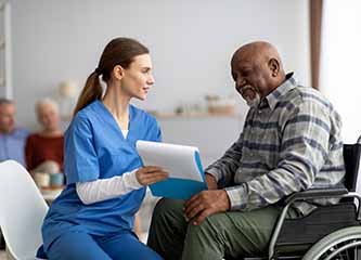 Pretty young woman in workwear nurse helping black senior man in weheelchair filling papers, cheerful elderly african american male patient signing documents at nursing home