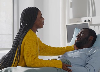 Afro-american wife visiting ill husband in hospital room. Sick african man patient resting in hospital bed and hugging girlfriend during visit in clinic