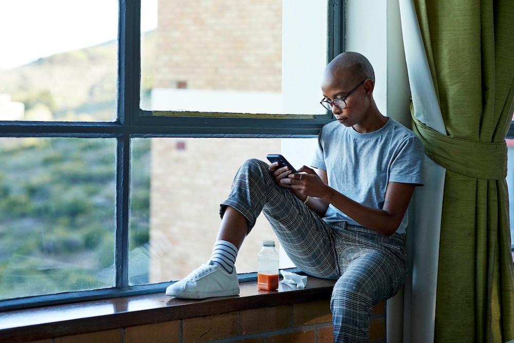 Completely bald female student sitting on window sill with juice while text messaging through smart phone in college cafeteria