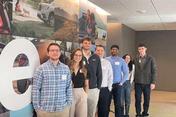 A group of male and female students pose in front of an image mural at Suburu of America.