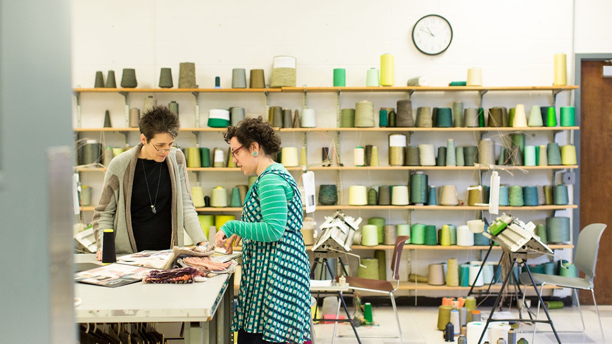 Two women discussing fabric samples in front of a shelving unit featuring an entire wall of thread spools