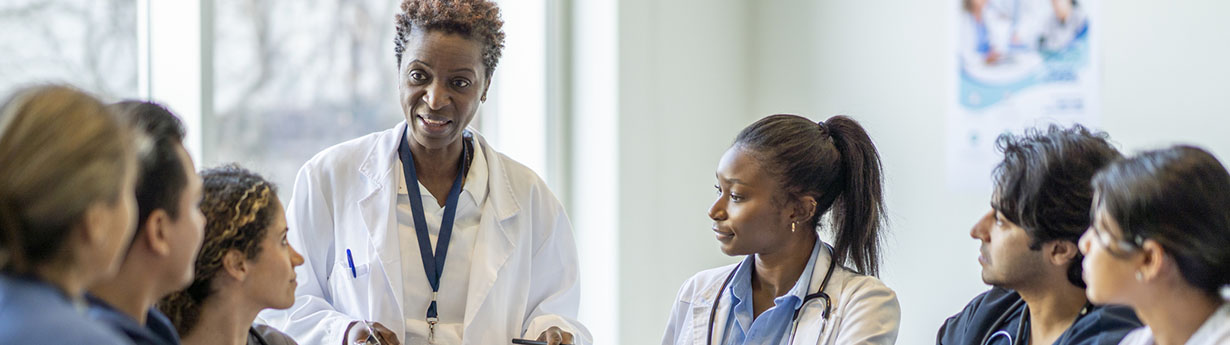 A small group of diverse nursing students sit around a boardroom table as they listen attentively to their teacher and lead doctor.  They are each dressed in medical scrubs and sitting with papers out in front of them.  The doctor is holding out a clipboard with a document on it as she reviews it with the group.