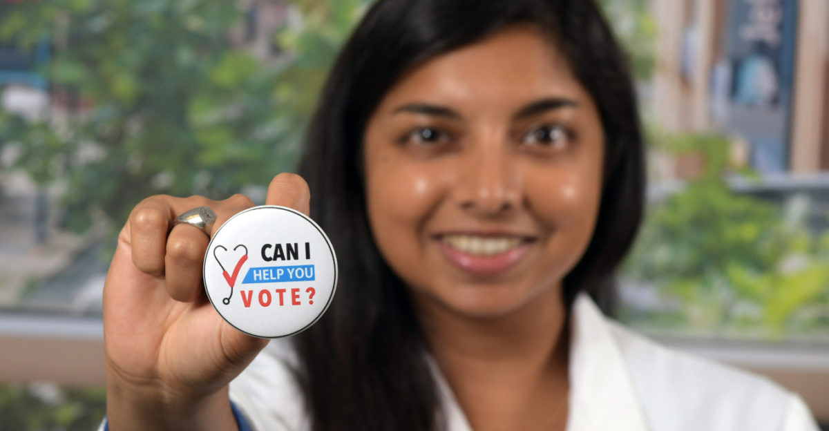 Medical student holds up a pin that says Can I Help You Vote?