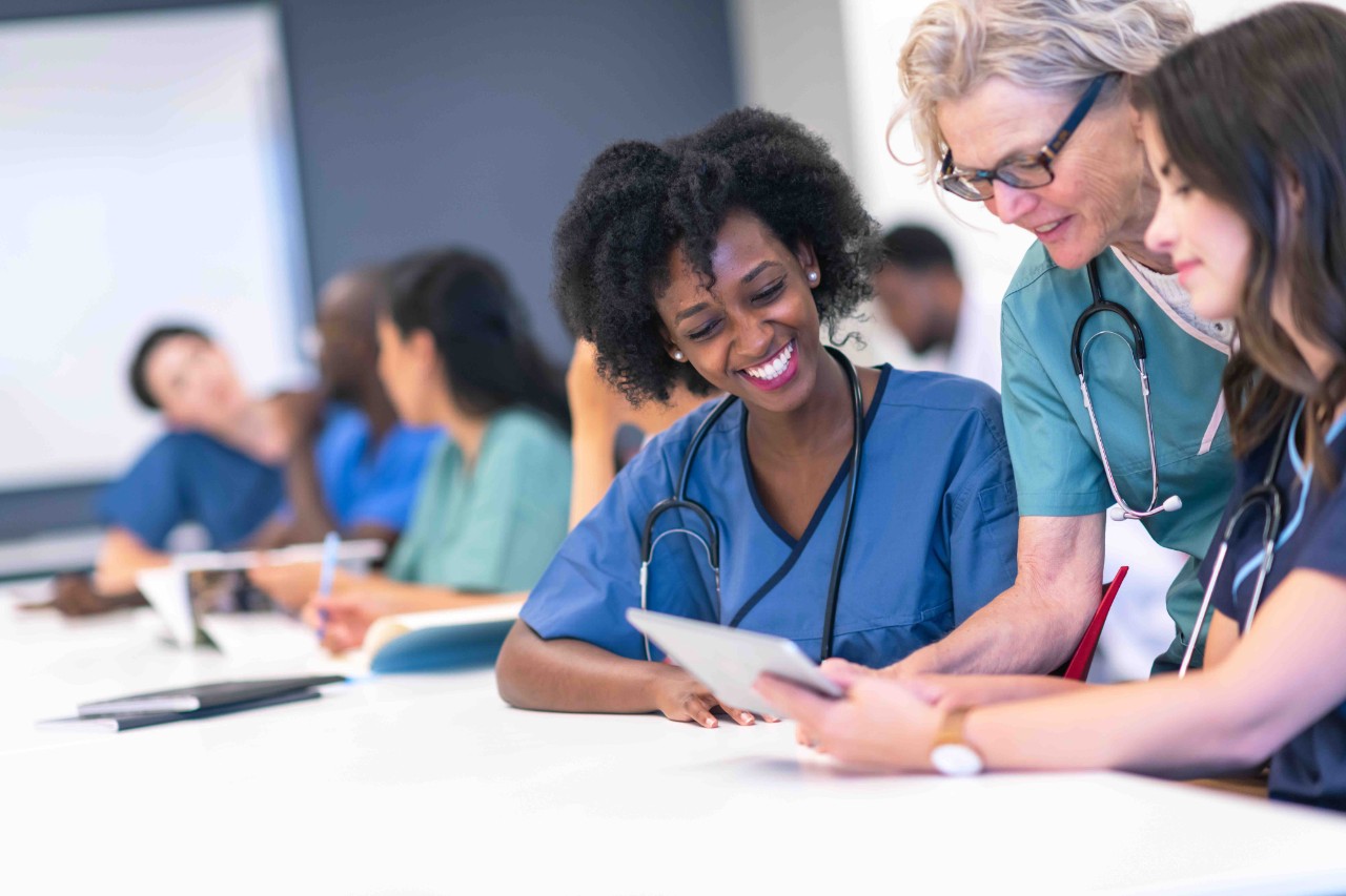 A group of nursing students are attending class together. They are seated at a long table in a classroom. The individuals are working in pairs on a project. The two women seated closest to the camera are being assisted by their female professor who is showing them something on a digital tablet.